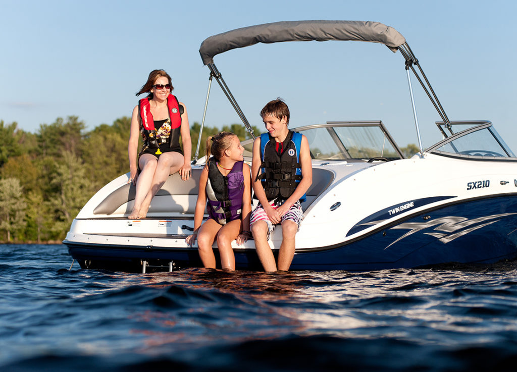 Kids and a woman sit at the back of a boat while wearing life jackets.