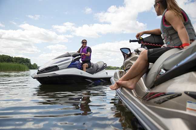 Two women wearing lifejackets on personal watercrafts, preseason boat maintenance concept.