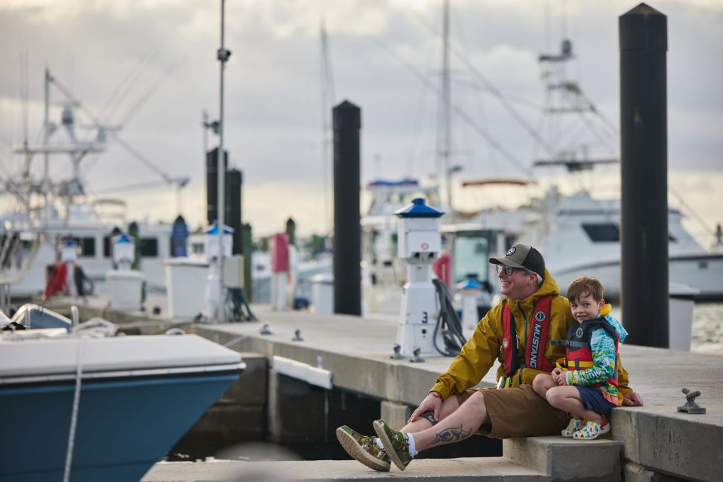 A man and boy sit on the dock while wearing life jackets.