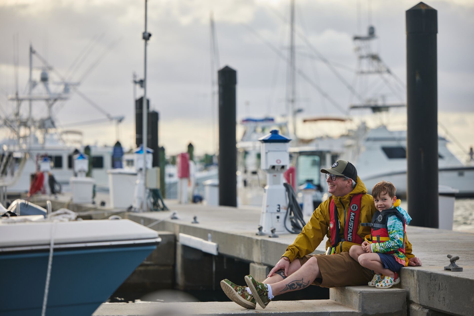 A smiling man and a boy sit on the dock of a marina while wearing life jackets.
