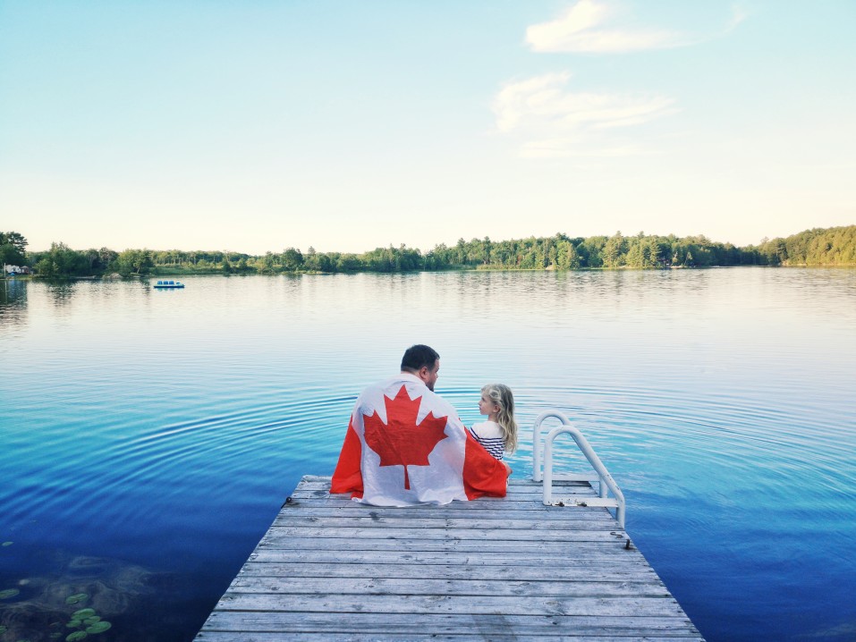 A man draped in a Canadian flag sits on a dock on a lake with a child, boating in Prince Edward Island concept. 