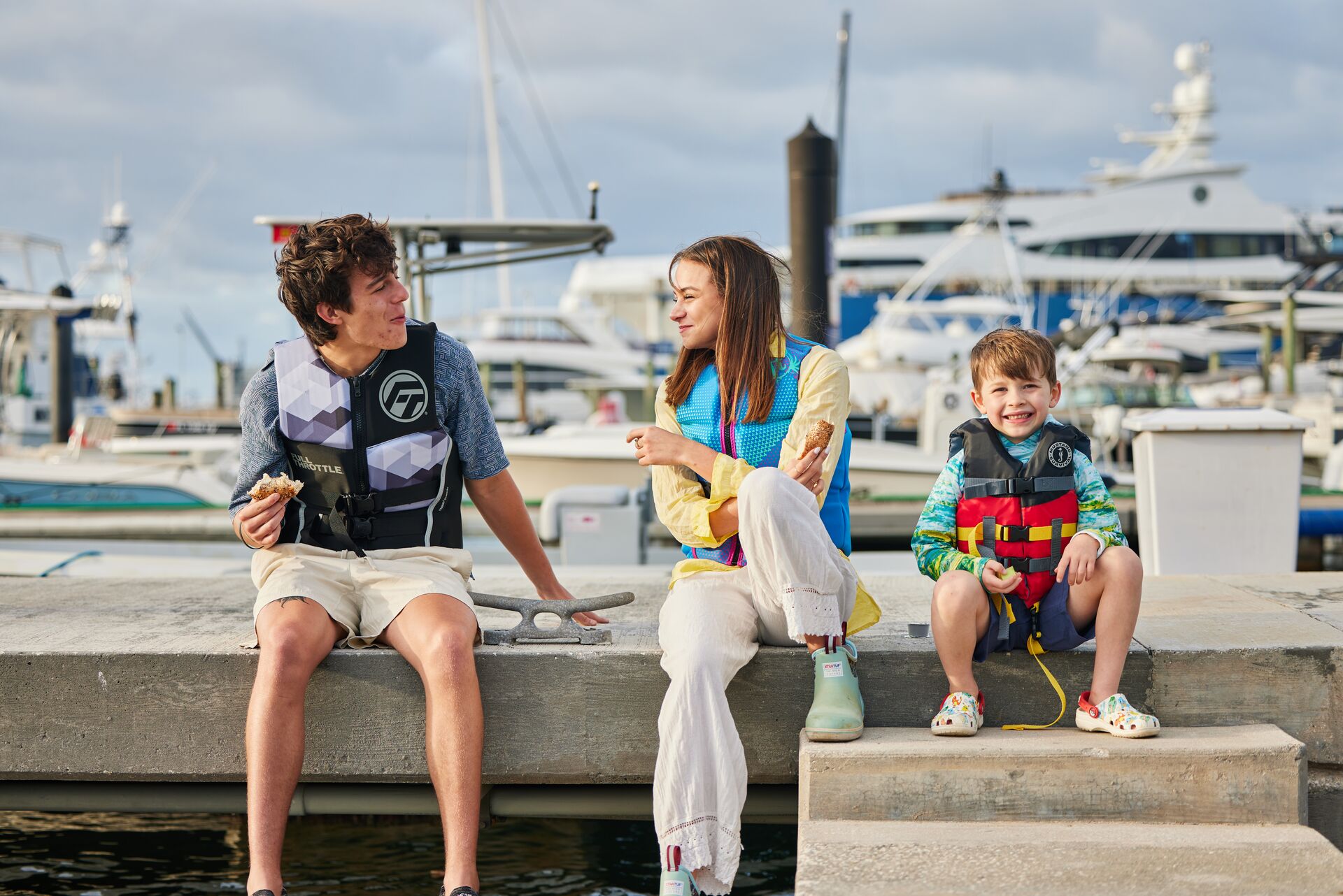 Three kids sitting on a dock while wearing life jackets.